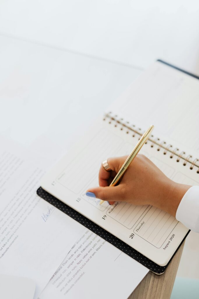 Personne qui écrit dans son agenda avec un crayon or sur une table avec des papiers.