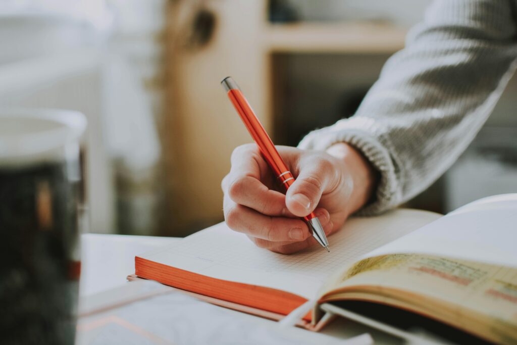 Image rapproché sur la mains d'une personne qui écrit dans un cahier avec un crayon rouge. Fond flouté.
